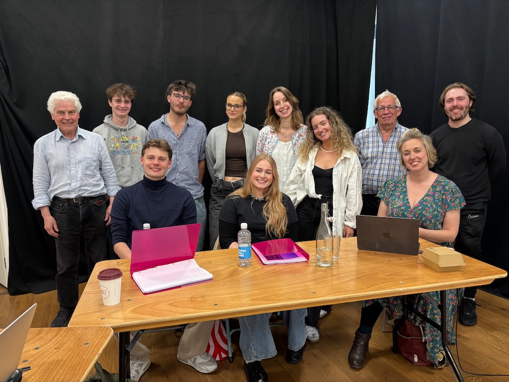 IMG_1094 Ian Grant and Chris Walker stand with eight young, white National Youth Music Theatre performers and Kate Golledge. There are four female students and four male. Two of the young performers and Kate sit in front of the others and behind a table. There are folders, a laptop, a glass and a plastic water bottle, takeaway coffee and food box on the table. Behind everyone are black curtains.