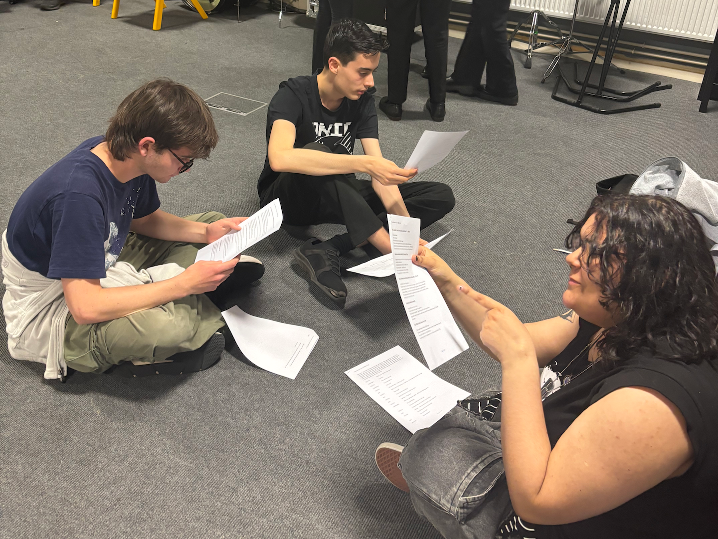 Three pupils sit on the floor of a rehearsal room reading scripts.