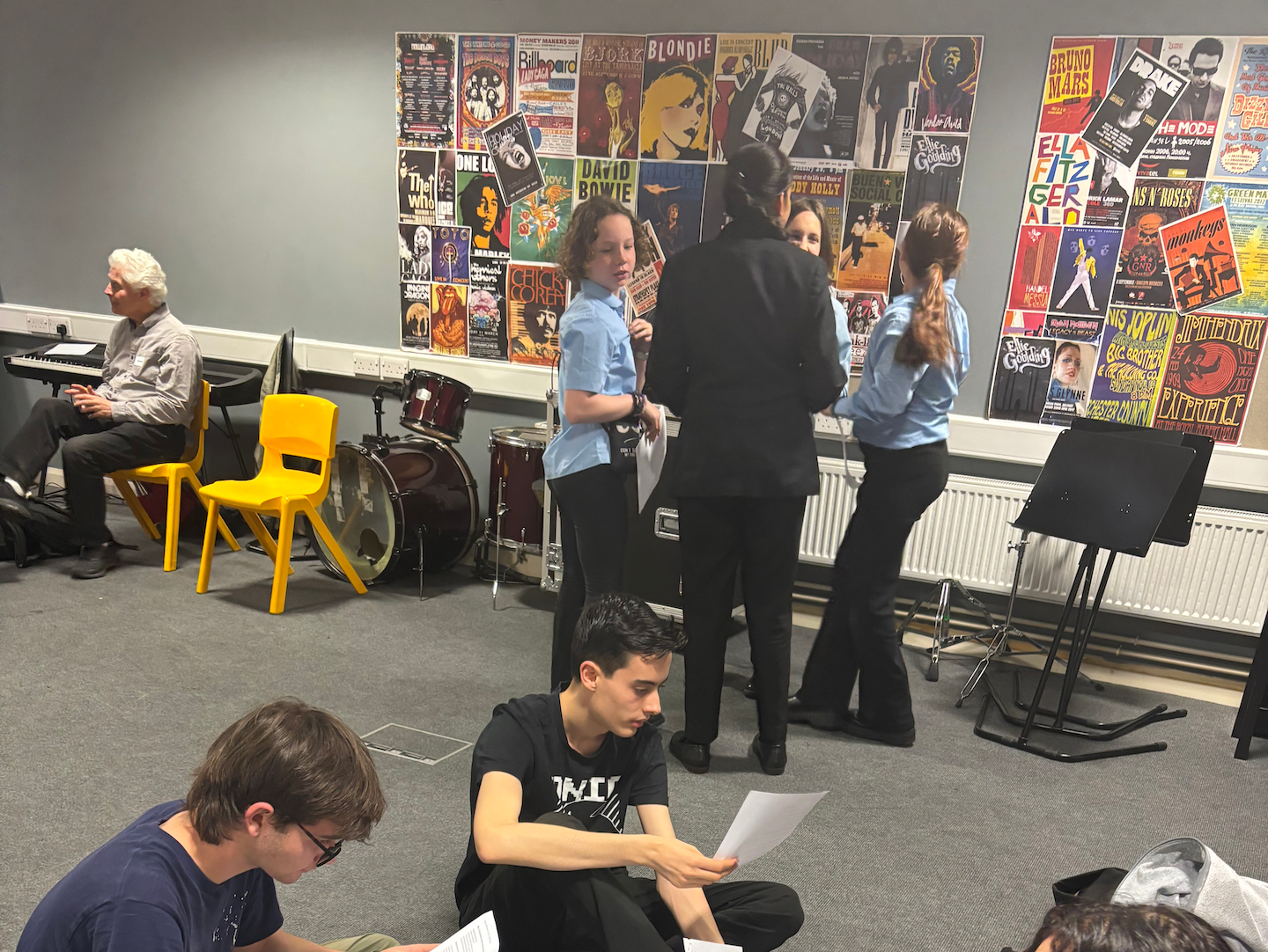 Three pupils sit on the floor of a rehearsal room reading scripts. Behind them is a group of four pupils also holding scripts and surrounded by music stands and a drum kit. There is a poster collage of album covers on the wall. Ian Grant sits on a yellow chair to the top left of the image.
