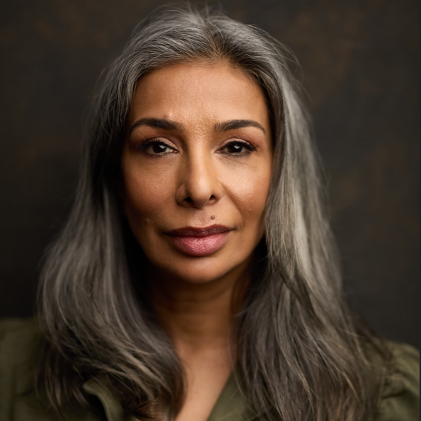 Shobna Gulati A headshot of Shobna Gulati, a middle-aged brown non-binary person with long dark grey hair. They have a mole above the left side of their lips and strong, manicured dark eyebrows. The background is dark grey and they are wearing an olive green shirt.