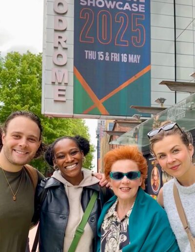 Jack Reitman, Cassiopeia Berkeley-Agyepong, Sophie-Louise Dann and Amy Parker pose in front of the Birmingham Hippodrome building sign. There is a BEAM showcase 2025 poster displaying on the screen above their heads.