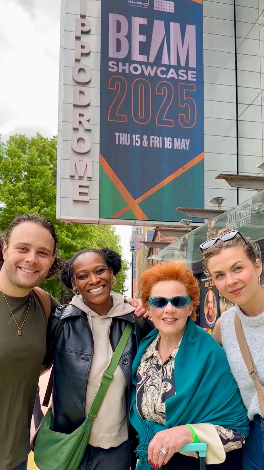 Jack Reitman, Cassiopeia Berkeley-Agyepong, Sophie-Louise Dann and Amy Parker pose in front of the Birmingham Hippodrome building sign. There is a BEAM showcase 2025 poster displaying on the screen above their heads.
