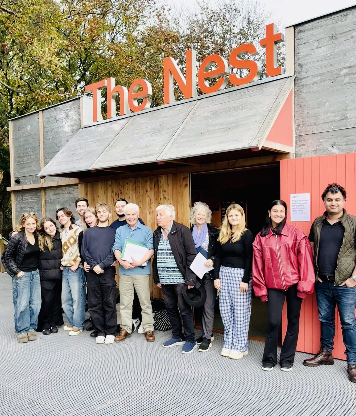 Ten students pose with Ian Grant, Christopher Walker and Meredith Braun in front of a wooden single-story building. The building name, The Nest, appears in big orange letters at the top of the building.