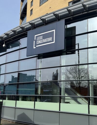 A curved glazed building with yellow brickwork above. A black sign over the windows reads LEEDS CONSERVATOIRE in white text with a broken rectangle around the words.