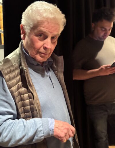 Ian Grant, an older white man with white hair, looks up into the camera from under his brow. He is wearing a pale blue shirt and pale blue zip-neck jumper with a puffed metallic gilet over the top. Behind him is a black curtain and an out of focus Josh Sood looks at his phone in the background.