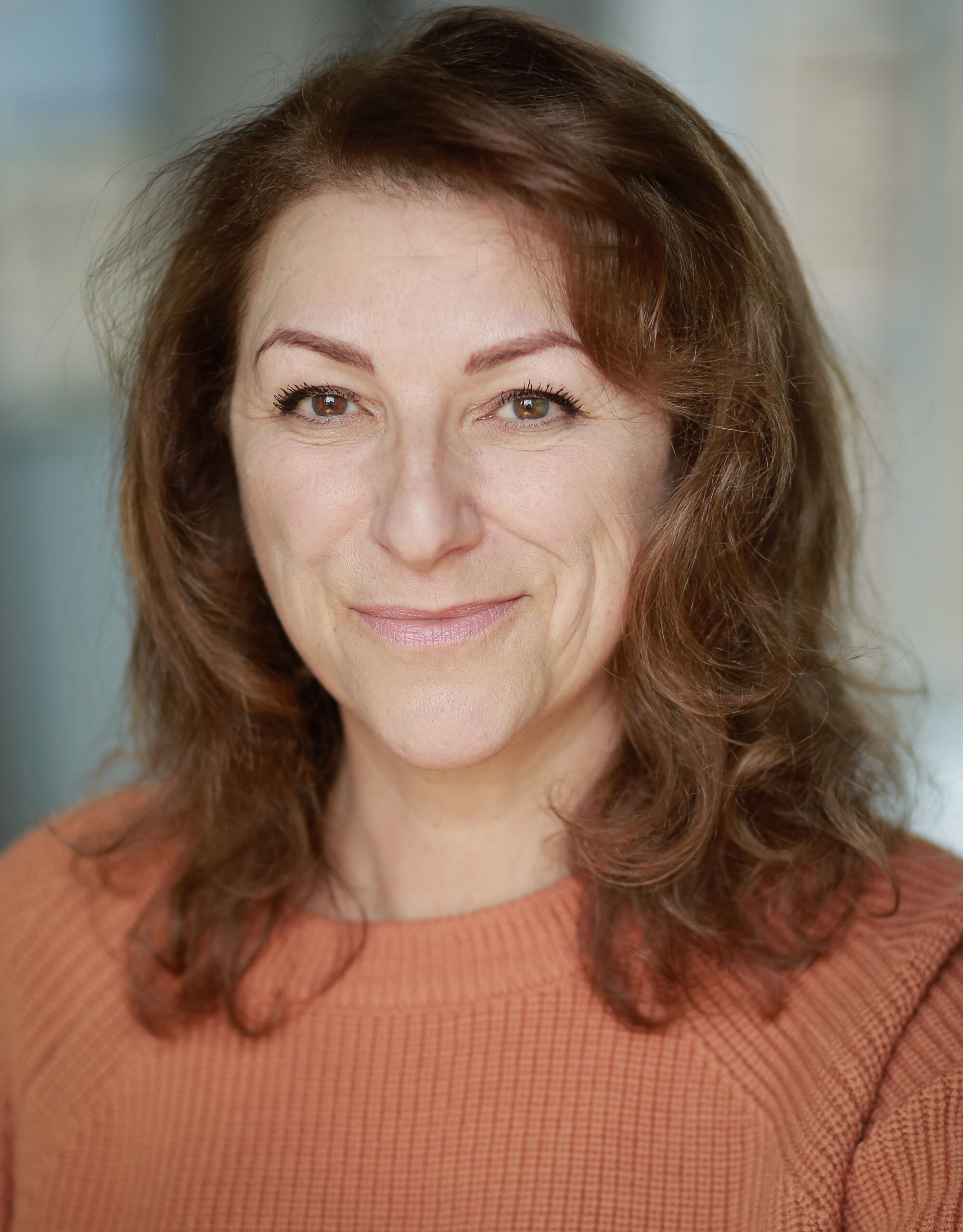 Headshot of Kim Ismay, a middle-aged white woman with brown eyes and light brown shoulder-length hair. She is wearing an orange-brown ribbed jumper and is smiling.