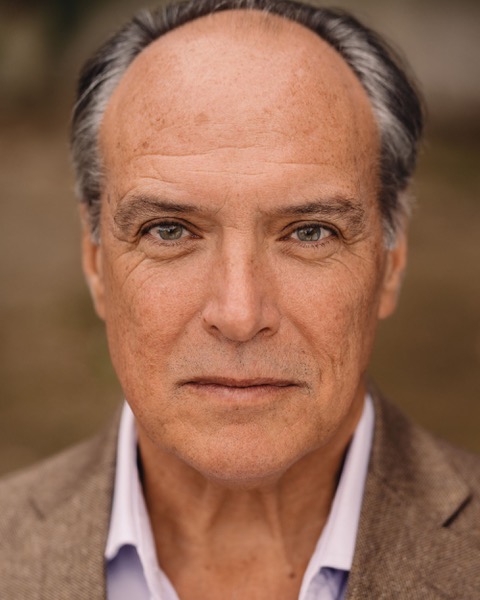 Headshot of Mark Carlisle. He is an older white man balding on top with dark grey hair to the back and sides of his head. He has defined eyebrows and wears a brown jacket over a lilac shirt.