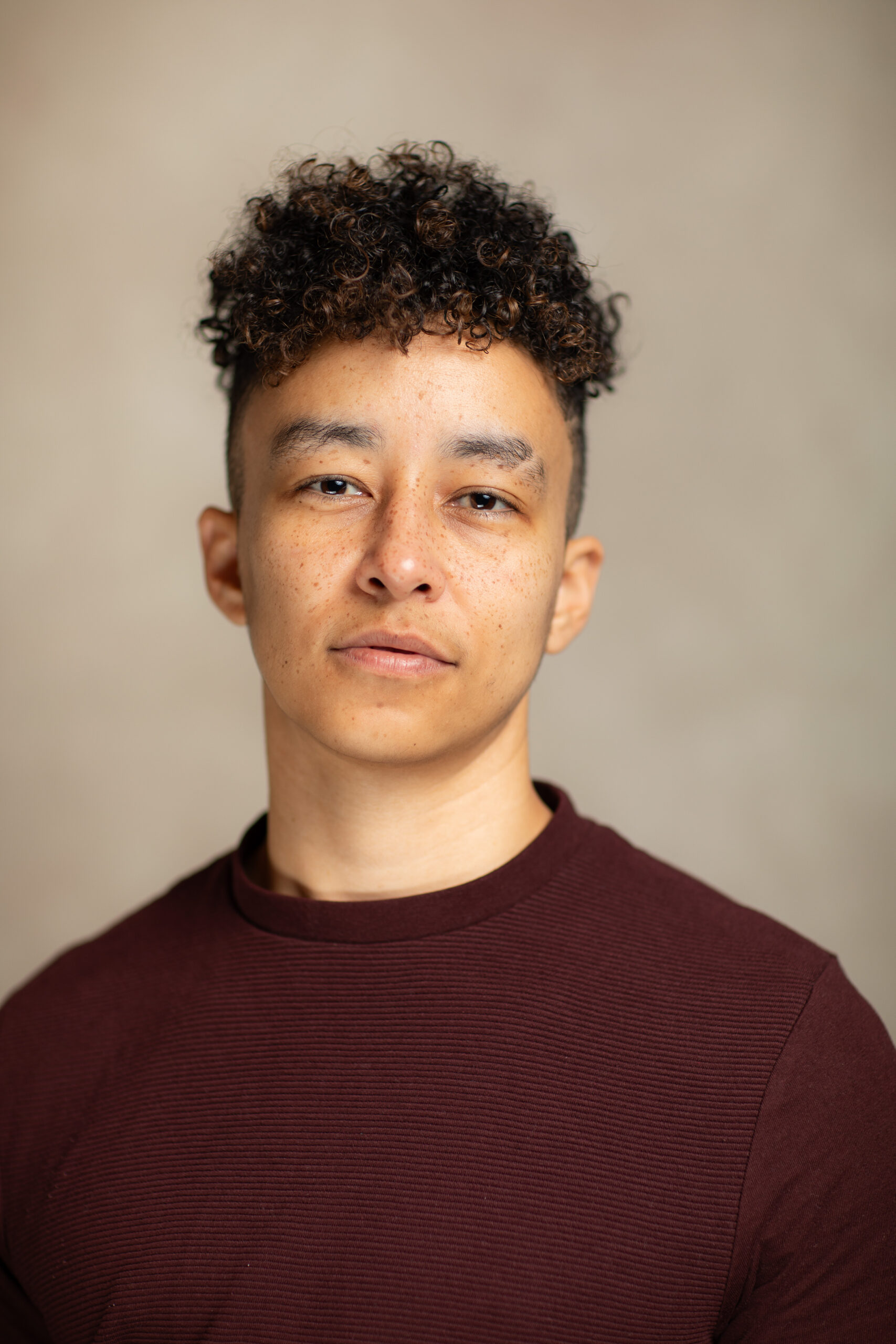 Headshot of Sé Carr. They are a brown actor with curly brown short hair buzzed above the ears. They have an eyebrow cut. They are wearing a maroon jumper.