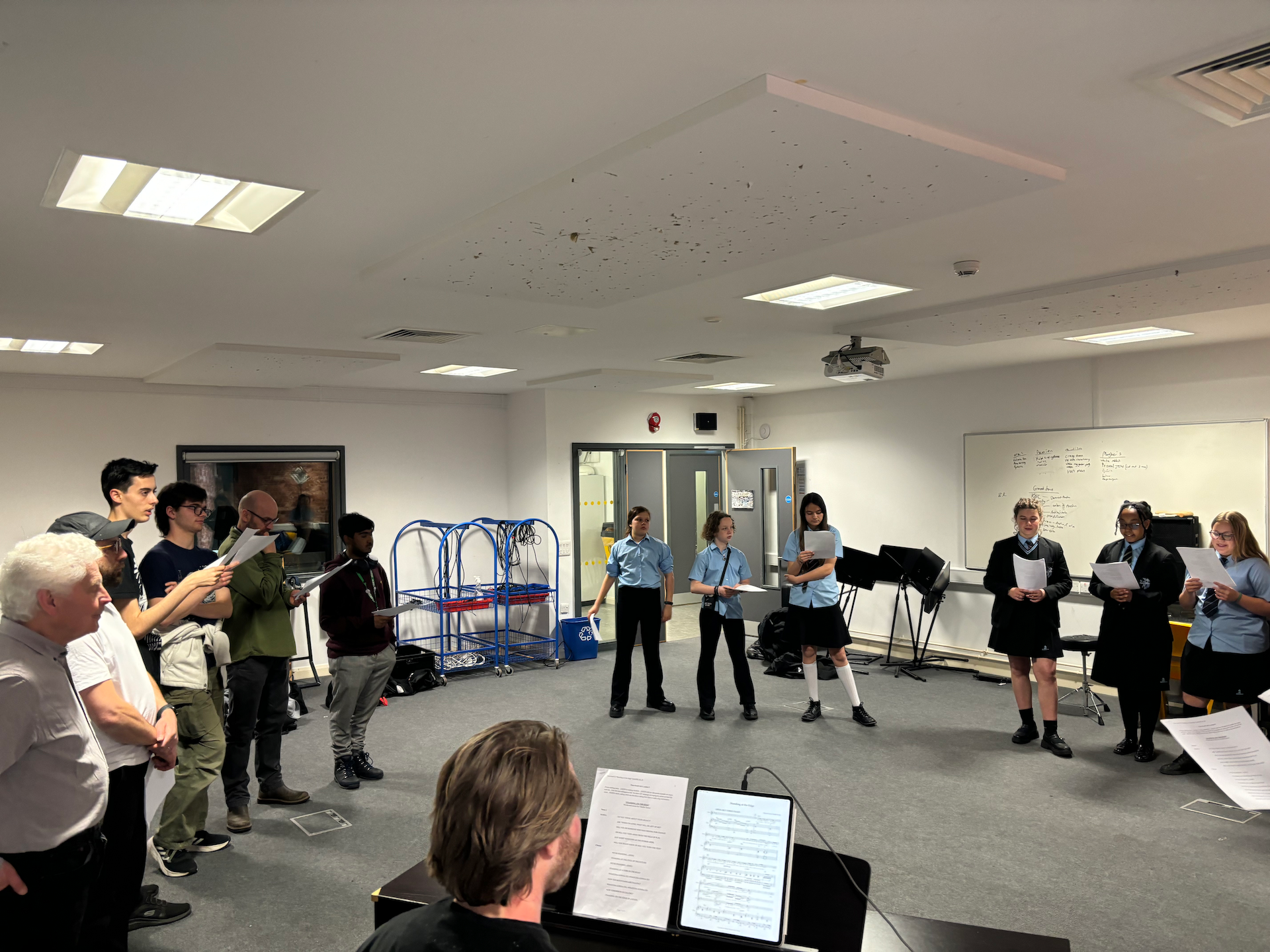 Ian Grant & Gavin Dent stand amidst a circle of pupils. Some are wearing school uniform and some are in their own clothes. Many of them hold scripts. Also in the circle is a piano with a pianist playing. They are all in a rehearsal room with grey walls and carpet. There is a whiteboard on the back wall with music stands in front of it.