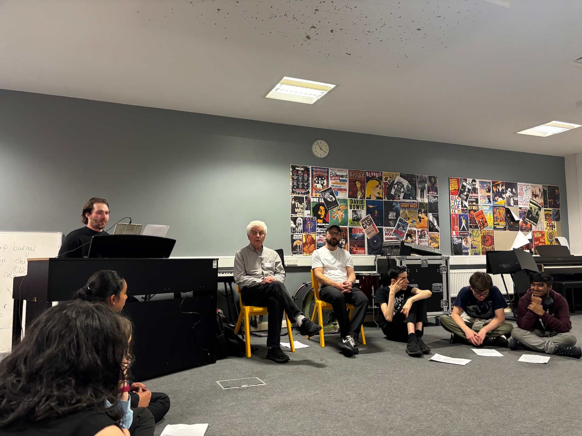 Ian Grant & Gavin Dent sit on yellow chairs amidst a circle of pupils sitting on the floor. Also in the circle is a piano with a pianist playing. They are all in a rehearsal room with grey walls and carpet. On the wall is a poster collage of album covers.