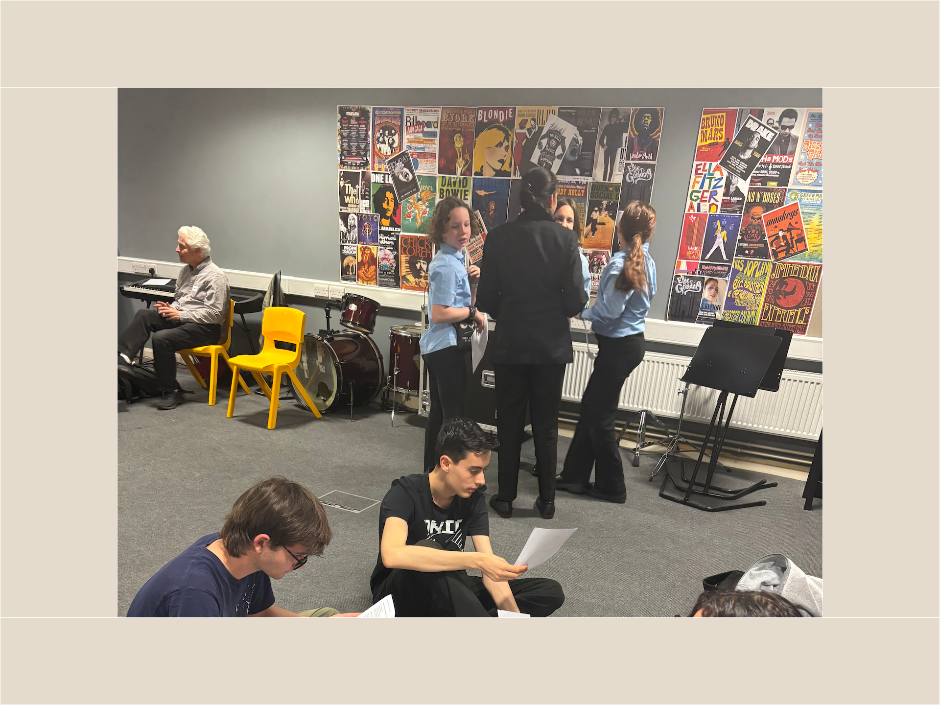 Three pupils sit on the floor of a rehearsal room reading scripts. Behind them is a group of four pupils also holding scripts and surrounded by music stands and a drum kit. There is a poster collage of album covers on the wall. Ian Grant sits on a yellow chair to the top left of the image.