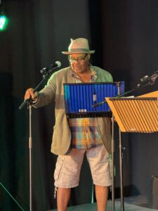 Clive Rowe wearing a trilby hat, shorts, bright check shirt and olive linen jacket. He is standing at a microphone on a stand in front of black curtains and lit by stage lights.
