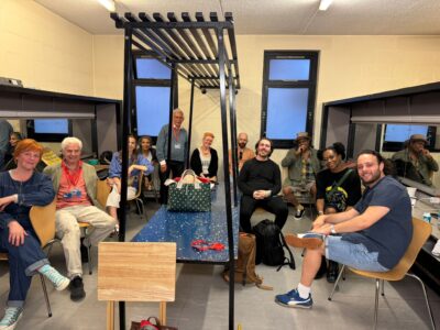 Sophie-Louise Dann, Ian Grant, Amy Parker, Shobna Gulati, Chris Walker, P Burton-Morgan, Gavin Dent, Tom Mitchell, Clive Rowe, Cassiopeia Berkeley-Agyepong & Jack Reitman sit on chairs around the edge of a rectangular dressing/changing room. In the middle is a table with overhead storage structure.