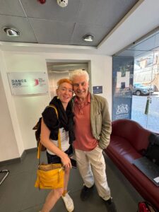 P Burton-Morgan & Ian Grant pose together in front of an elevator in a lobby of Birmingham Hippodrome. P has orange hair with shaved sides and wears a black scarf, a white top and denim shorts with white trainers. They have a yellow leather handbag slung over their shoulder. Ian has white hair and wears a khaki jacket over a salmon polo shirt and white trousers with black shoes.