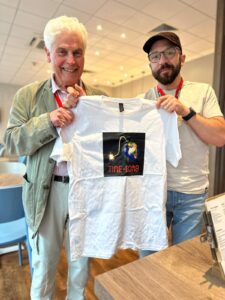Ian Grant and Gavin Dent hold up a white t-shirt with the Time-bomb logo printed on it: The words 'TIME-BOMB' are in red letters that look like those of a digital clock. Below this is a lit fuse leading to a bomb on the right-hand side of the image. The bomb is made to look like planet earth. The background is black. Ian Grant has white hair and is wearing a khaki jacket. Gavin Dent is wearing a brown cap, large glasses and has a dark beard.