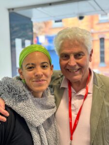 EM Williams & Ian Grant pose together. EM Williams wears a bright green durag and a grey fleece scarf. They have a septum piercing and an eyebrow slit. Ian Grant has short white hair and is wearing a pale pink polo shirt and a khaki jacket with a red lanyard round his neck.
