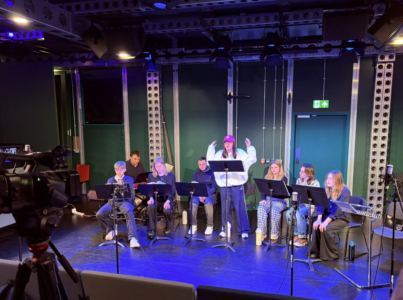 Seven students sit to either side of Serena Chahal who stands wearing a red cap. They are in front of music stands on the stage area of a studio theatre with green walls. In the background there is a pianist sitting at a red keyboard. There is a camera on a tripod in the left foreground of the image.
